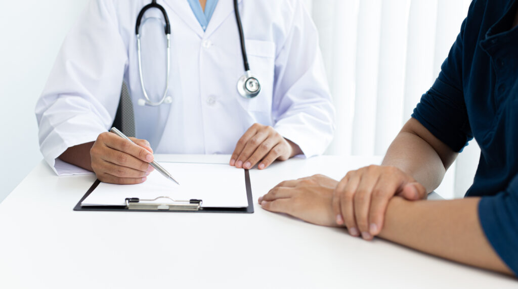 Um médico apontando uma caneta para sua prancheta e explicando para seu paciente os remédios que ele vai tomar.